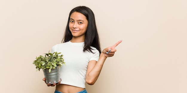 Mujer asiática joven que sostiene una planta que sonríe y que señala a un lado, mostrando algo en el espacio en blanco.