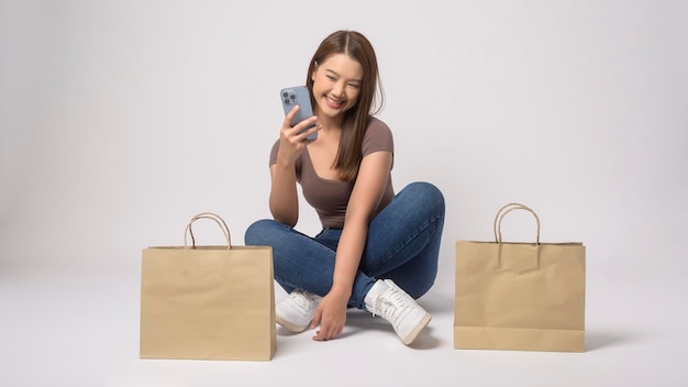 Mujer asiática joven que sostiene el smartphone y el bolso de compras sobre el fondo blanco