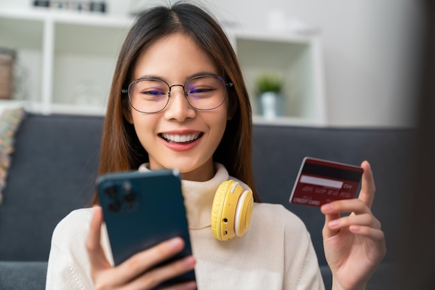 Mujer asiática joven que sostiene la tarjeta de crédito y que usa el teléfono inteligente para comprar en línea con pago en banca por Internet.