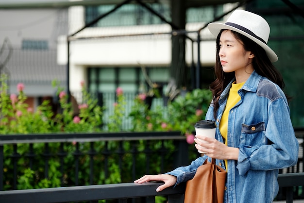 Mujer asiática joven que sostiene una taza de café de papel para llevar mientras que se coloca al aire libre fondo de la ciudad