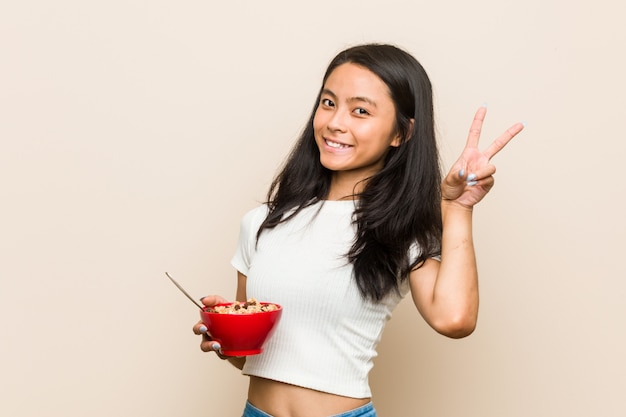La mujer asiática joven que sostiene un tazón de cereales alegre y despreocupado que muestra un símbolo de paz con los dedos.