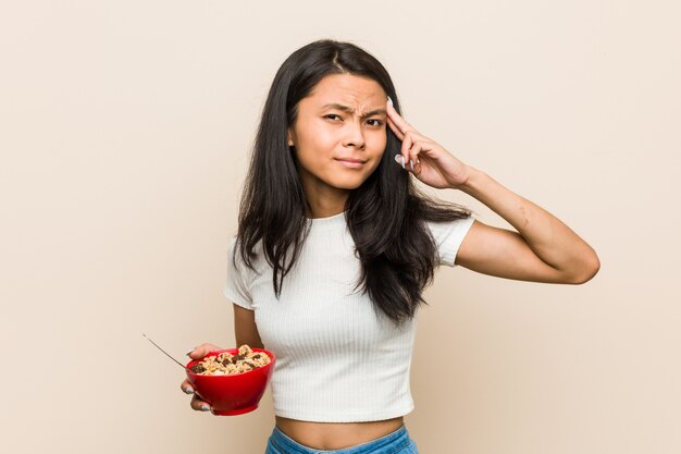 Mujer asiática joven que sostiene un tazón de cereales que muestra un gesto de decepción con el índice.