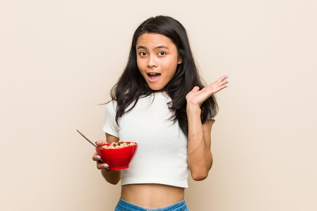 Mujer asiática joven que sostiene un tazón de cereales sorprendido y conmocionado.