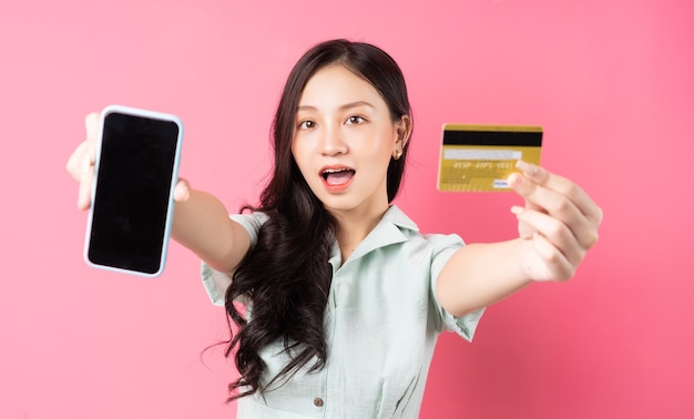 Mujer asiática joven que sostiene un teléfono móvil mientras sostiene una tarjeta bancaria en su mano en rosa