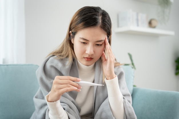 Mujer asiática joven que tiene fiebre alta mientras controla la temperatura corporal estornudos femeninos y secreción nasal con gripe estacional termómetro digital alérgico virus coronavirus enfermedad respiratoria