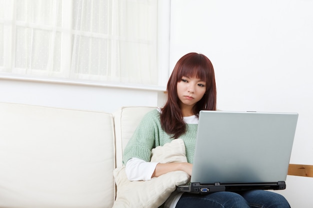 Mujer asiática joven que usa la computadora portátil en casa en el sofá.