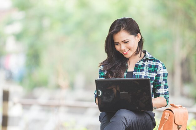 Mujer asiática joven que usa la computadora portátil mientras está sentado en el parque