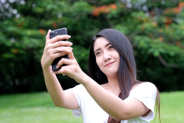 Mujer asiática joven que usa el smartphone para selfie su retrato en el fondo del parque de naturaleza