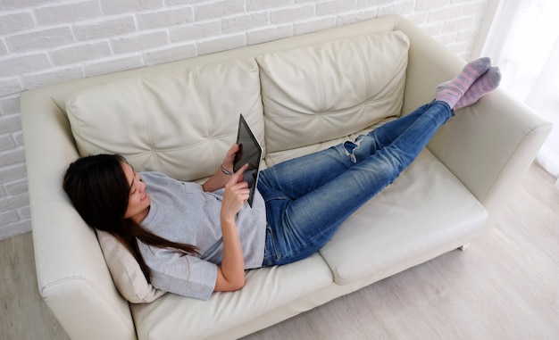 Mujer asiática joven que usa la tableta con emoción sonriente, feliz y relajada acostado en el sofá