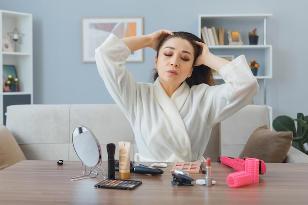 mujer asiática joven relajada con toalla en albornoz sentada en el tocador en el interior de casa tocando su cabello haciendo masaje de cabeza haciendo rutina de maquillaje matutino