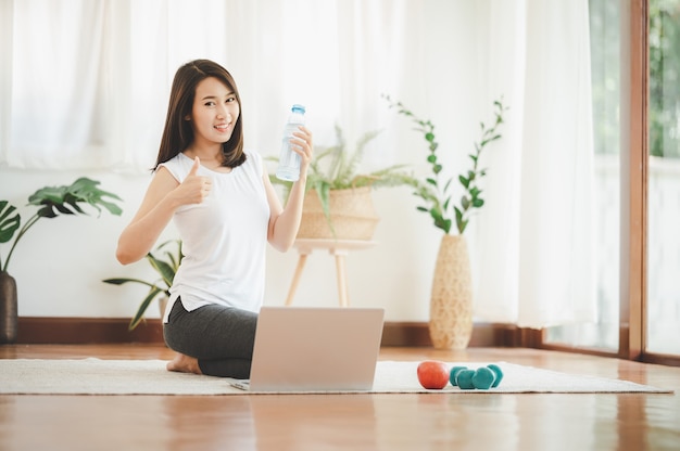 Mujer asiática joven sana que muestra el pulgar hacia arriba mientras sostiene la botella de agua lista para usar la computadora portátil para practicar yoga en línea o hacer ejercicio en casa