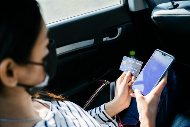 Mujer asiática joven sentada en el asiento trasero del coche y usando el móvil y sosteniendo la tarjeta de crédito en las manos.