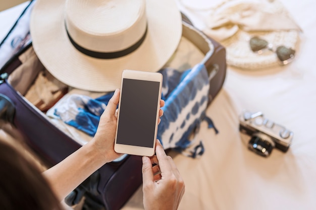 Mujer asiática joven sentada en la cama con teléfono inteligente y empacando su maleta preparándose para viajar en vacaciones de verano