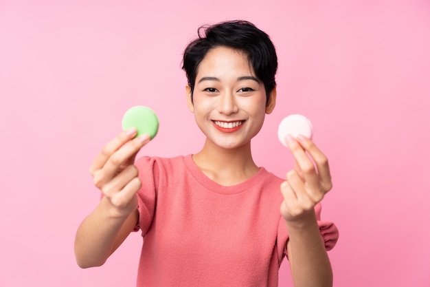 Mujer asiática joven sobre la pared rosada aislada que ofrece macarons franceses coloridos