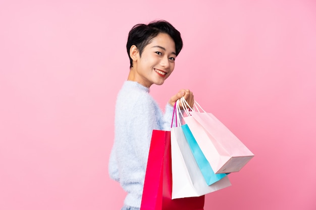 Mujer asiática joven sobre la pared rosada aislada que sostiene bolsos de compras y que sonríe