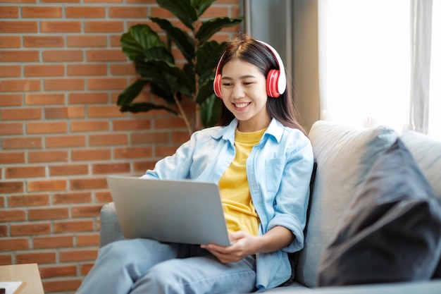 Mujer asiática joven sonriendo escuchando música usando una computadora portátil mientras está sentada en el sofá en casa