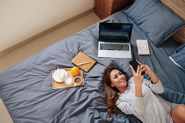 Mujer asiática joven sonriente atractiva que se relaja en la cama en casa con la bandeja del desayuno y que revisa las redes sociales en su teléfono inteligente