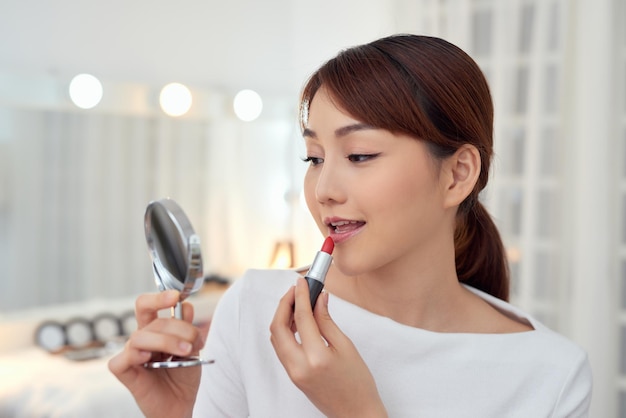 Mujer asiática joven sonriente que aplica el lápiz labial y que mira al espejo