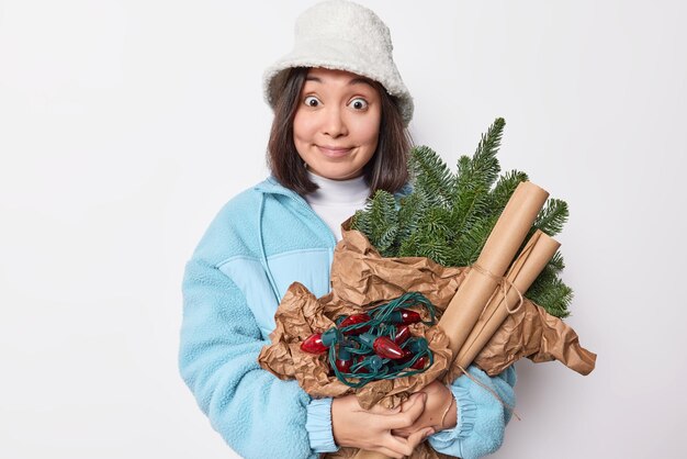 Mujer asiática joven sorprendida viste panamá de invierno blanco y chaqueta azul sostiene guirnaldas de ramas de abeto verde y papel espera para poses de Año Nuevo en el interior. Concepto de celebración y vacaciones de personas.