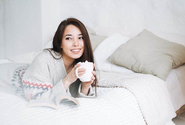 Mujer asiática joven en suéter de punto acogedor con taza de cacao con malvavisco en el dormitorio en casa