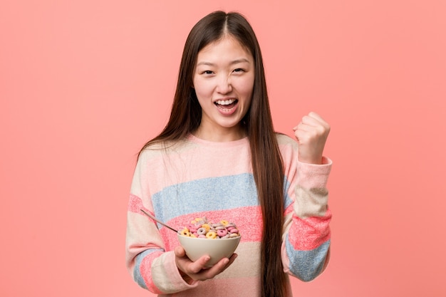 Mujer asiática joven con un tazón de cereal que anima despreocupado y emocionado. Concepto de victoria
