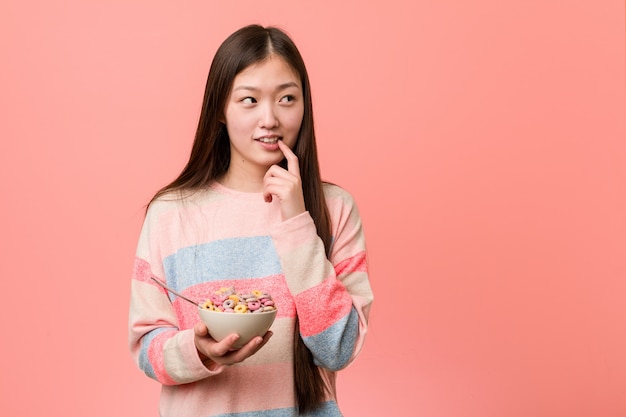 La mujer asiática joven con un tazón de cereal relajado pensando en algo mirando un espacio de copia.