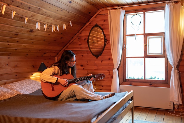 Mujer asiática joven tocando la guitarra acústica mientras se sienta en la cama en el hogar acogedor Concepto de afición