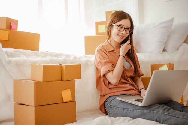 Mujer asiática joven trabaja desde casa hablando con los clientes para vender productos en línea y entregarlos al cliente. Propietario de negocio, PYME, envío, trabajo desde casa (FMH), autónomo, concepto de puesta en marcha.