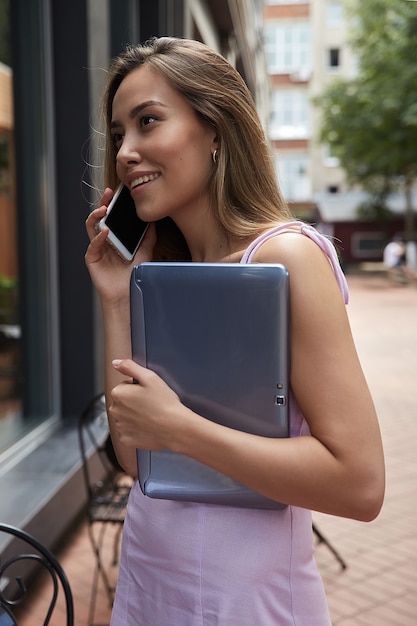 Mujer asiática joven en vestido de pie al aire libre con ordenador portátil llamando por teléfono inteligente