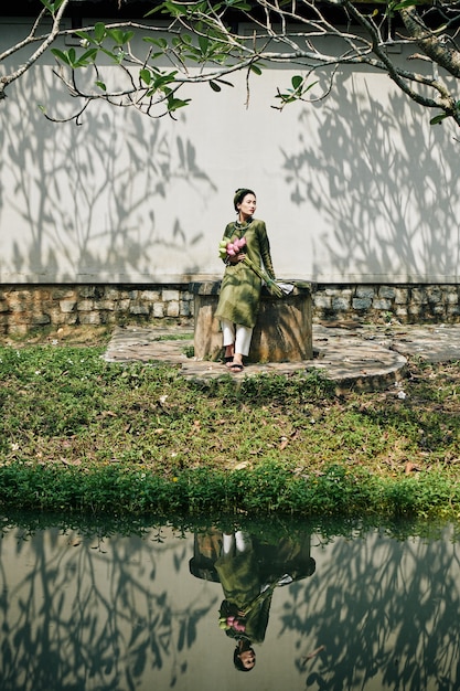Mujer asiática joven en vestido tradicional vietnamita ao dai sentado cerca de un pequeño estanque con flores de loto en las manos