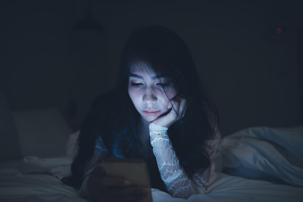 Mujer asiática jugando en el teléfono inteligente en la cama por la nocheGente tailandesaAdicto a las redes sociales