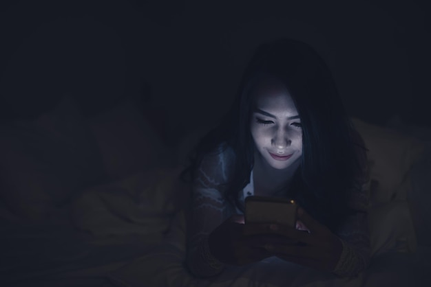 Mujer asiática jugando en el teléfono inteligente en la cama por la nocheGente tailandesaAdicto a las redes sociales