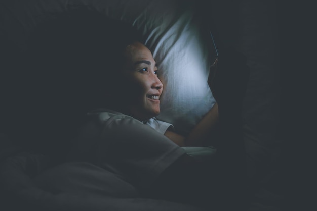 Mujer asiática jugando en el teléfono inteligente en la cama por la nocheGente tailandesaAdicto a las redes sociales