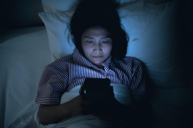 Mujer asiática jugando en el teléfono inteligente en la cama por la nocheGente tailandesaAdicto a las redes sociales