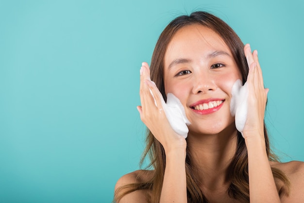 Mujer asiática lavado de cara exfoliación jabón exfoliante con limpieza de cuidado de la piel Retrato hermosa sonrisa femenina aplica limpiador espumoso piel sana Estudio aislado sobre fondo azul rutina diaria de cuidado de la piel