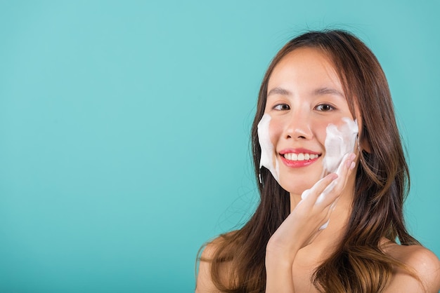 Mujer asiática lavado de cara exfoliar espuma de jabón exfoliante con limpieza de cuidado de la piel, retrato hermosa sonrisa femenina aplica limpiador de espuma piel sana, estudio aislado sobre fondo azul, cuidado de la piel de belleza