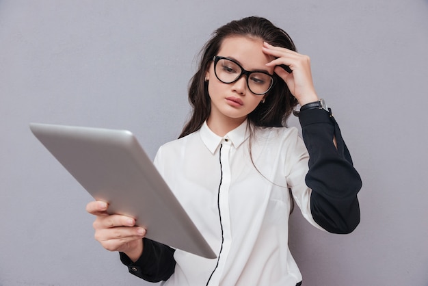 Mujer asiática linda joven en vidrios que leen en la computadora de la tableta. Centrarse en el modelo aislado en gris