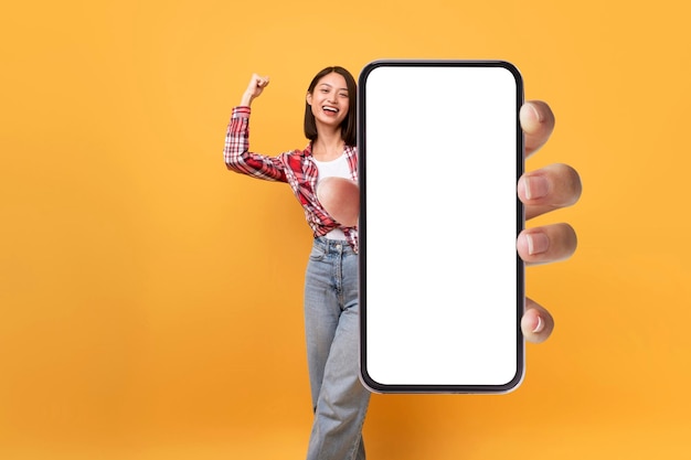 Mujer asiática llena de alegría que muestra una gran pantalla de teléfono inteligente y sacude el puño gran oferta móvil y espacio para su anuncio