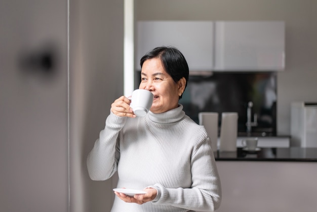 Mujer asiática madura tomando café cerca de la ventana por la mañana, feliz y sonriente, pensamiento positivo, concepto de seguro médico senior