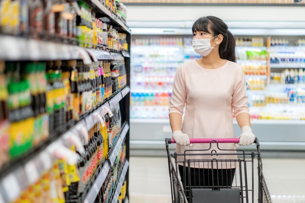 Mujer asiática con máscara higiénica y guante de goma con carrito de compras en el supermercado y buscando salsa o saborizante para comprar durante el brote de covid-19 para prepararse para una cuarentena pandémica