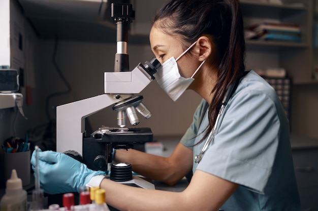 Mujer asiática con máscara médica mira al microscopio en la mesa en el hospital