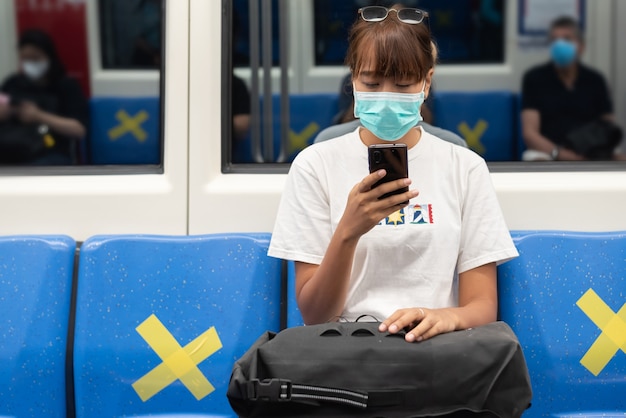 Mujer asiática con mascarilla quirúrgica se siente cansada usar smartphone sentado en el asiento azul en el metro