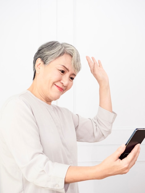 Mujer asiática mayor madura feliz que sostiene el teléfono inteligente usando aplicaciones móviles en línea.