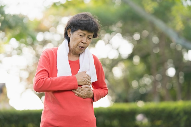 La mujer asiática mayor sufre de dolor del ejercicio. Mujer mayor que tiene un ataque al corazón después del ejercicio en el parque. Concepto de salud
