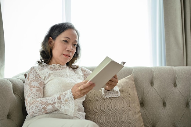 Una mujer asiática de mediana edad se concentra en leer un libro en su sala de estar