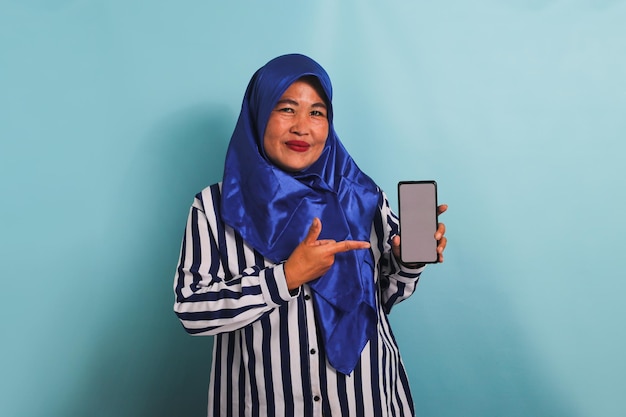 Una mujer asiática de mediana edad con hijab está apuntando con el dedo a su teléfono aislado sobre un fondo azul