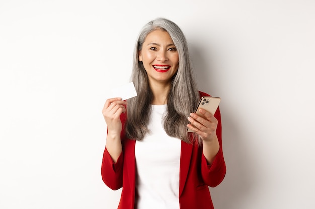 Mujer asiática de mediana edad con tarjeta de crédito de plástico y un teléfono inteligente para comprar en línea, de pie sobre fondo blanco.