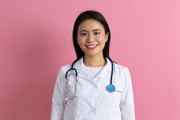 Mujer asiática médico en bata médica blanca con estetoscopio contra el retrato de fondo rosa