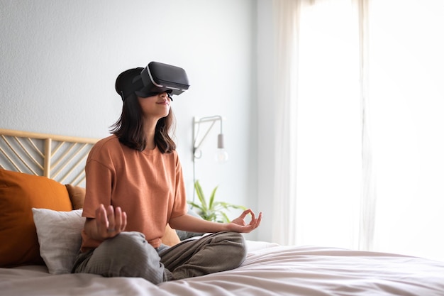 Mujer asiática medita con una aplicación de experiencia VR en la cama haciendo meditación usando gafas de realidad virtual