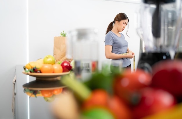 Mujer asiática midiendo su cintura con preocupación y emoción nerviosa con frutas y verduras borrosas en primer plano Vistiendo ropa deportiva Imagen con espacio de copia